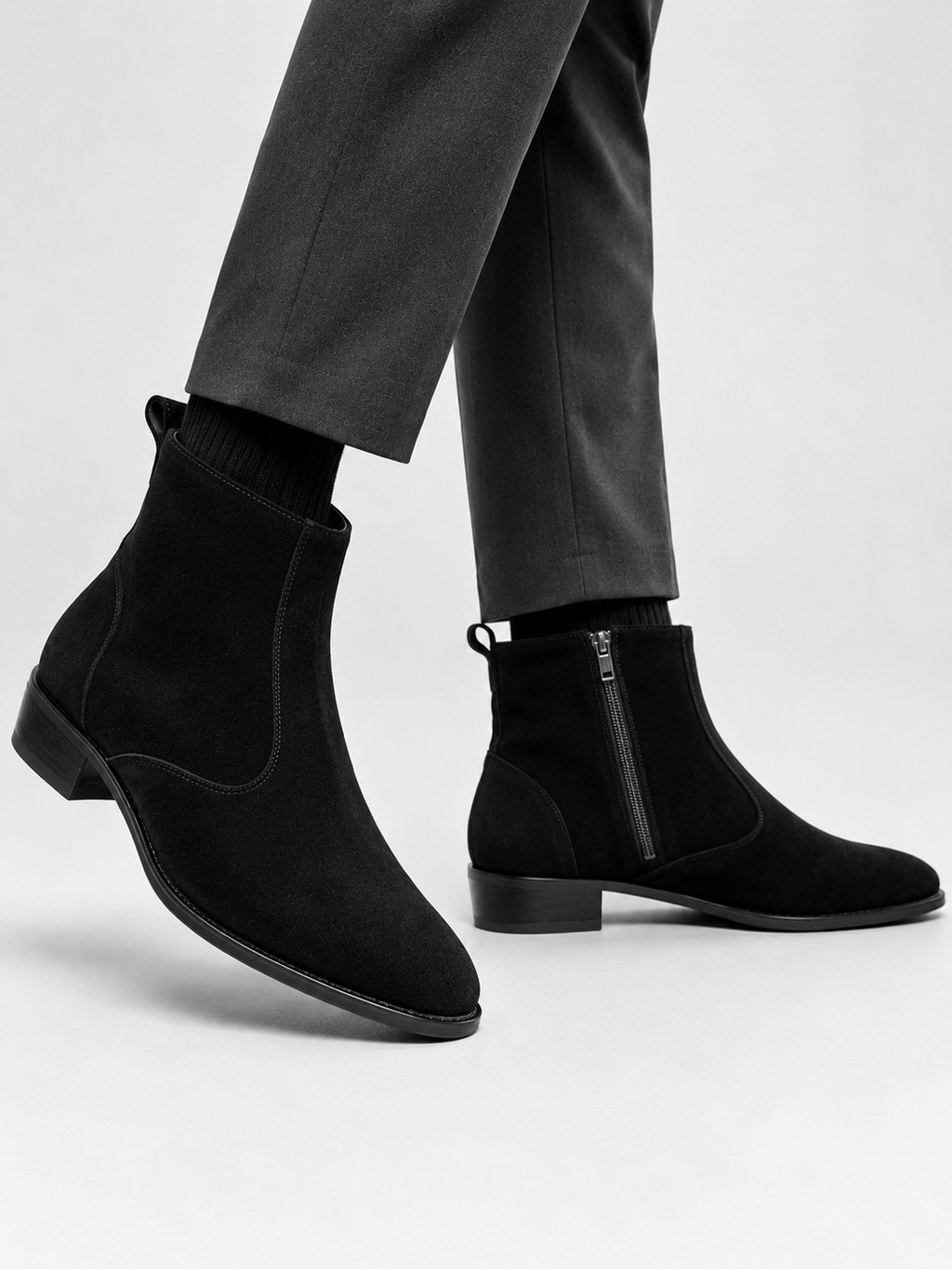Black suede Chelsea-style ankle boots with side zippers, shown on a model wearing dark gray trousers against a light gray studio background.