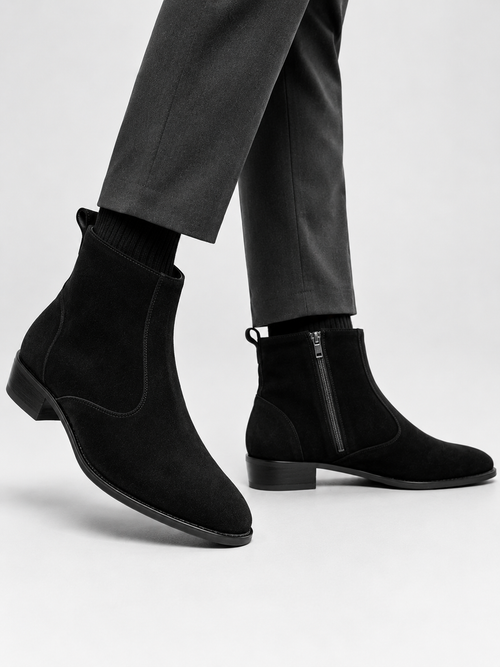Black suede Chelsea-style ankle boots with side zippers, shown on a model wearing dark gray trousers against a light gray studio background.