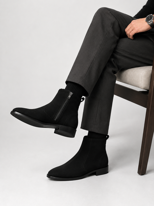 A person seated on a chair wearing black ankle boots with side zippers, black socks, and dark charcoal trousers against a light gray studio background.