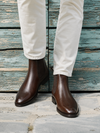 A person wearing dark brown leather Chelsea boots with elastic side panels and white trousers, standing against a weathered blue-green wooden plank wall.