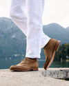 A person wearing white trousers and tan suede lace-up shoes, standing on a concrete ledge with a scenic mountain and lake background.