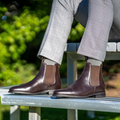 Person wearing brown leather Chelsea boots and gray trousers sitting on a bench with greenery in the background.