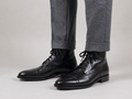 Premium black leather cap-toe lace-up dress boots for men paired with grey tailored trousers on a minimalist studio background.