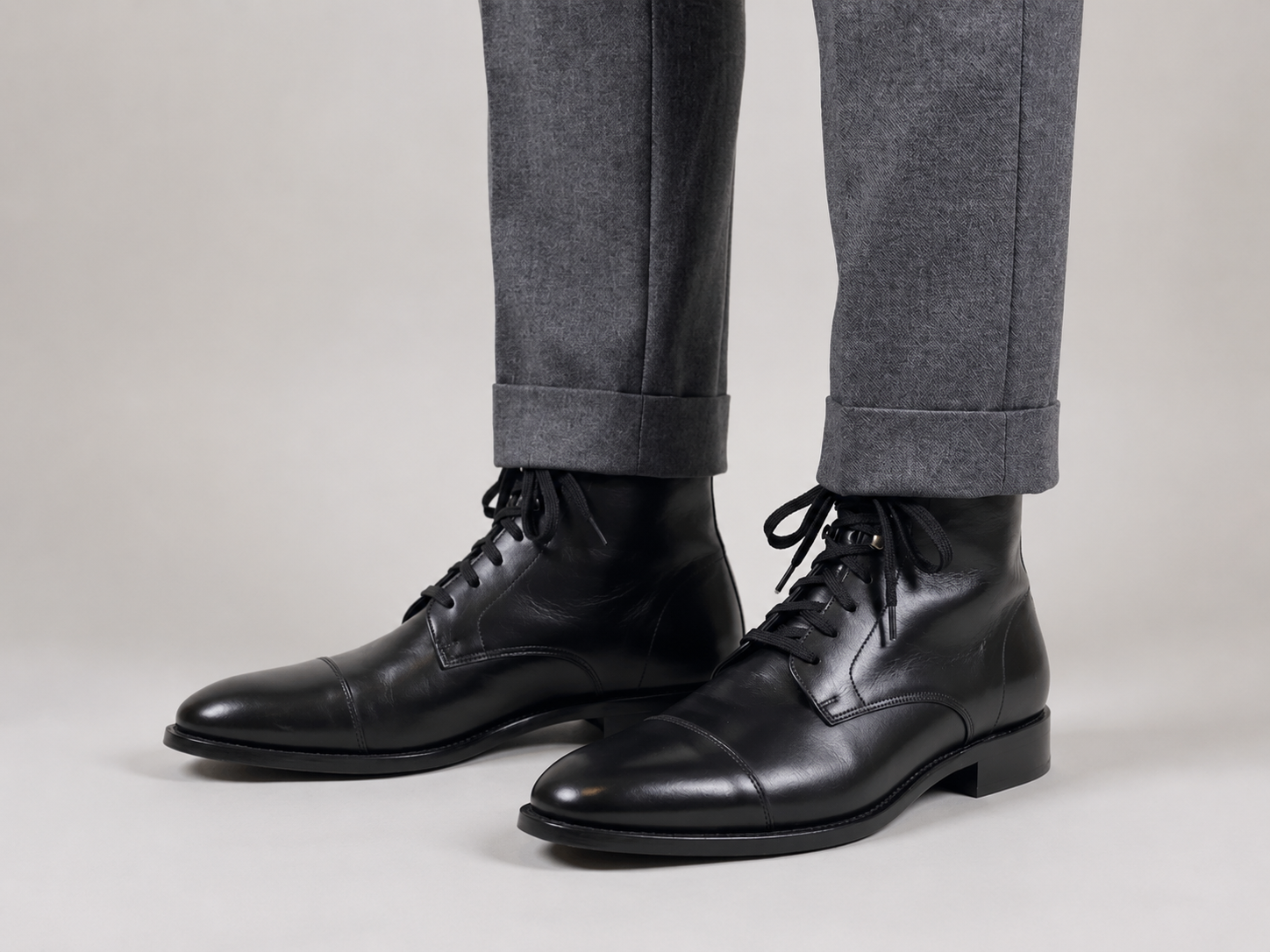 Premium black leather cap-toe lace-up dress boots for men paired with grey tailored trousers on a minimalist studio background.