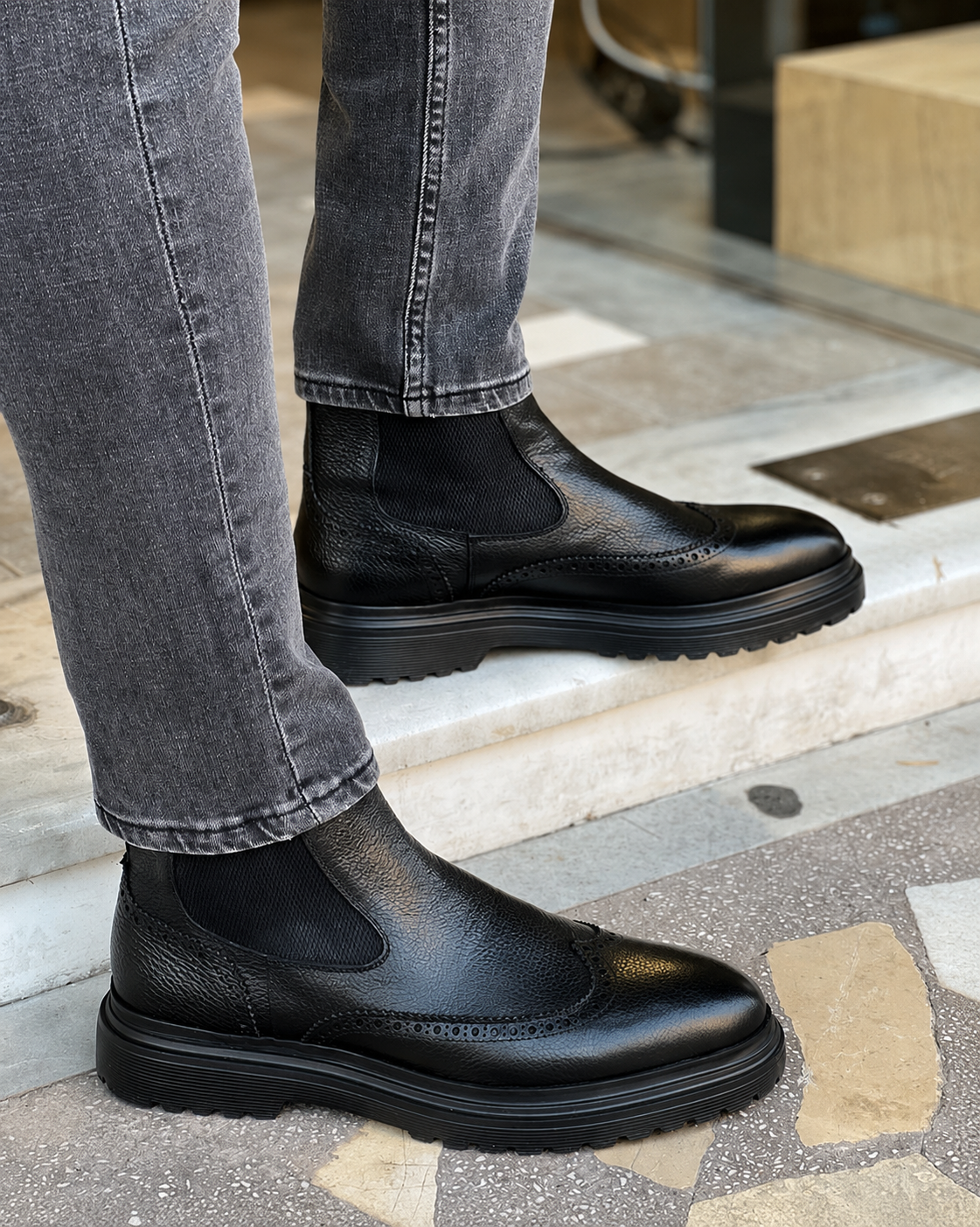 Black leather Chelsea boots worn with gray jeans on a stone pavement.