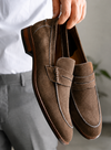 Brown suede loafers held by a person wearing a white shirt and gray pants.
