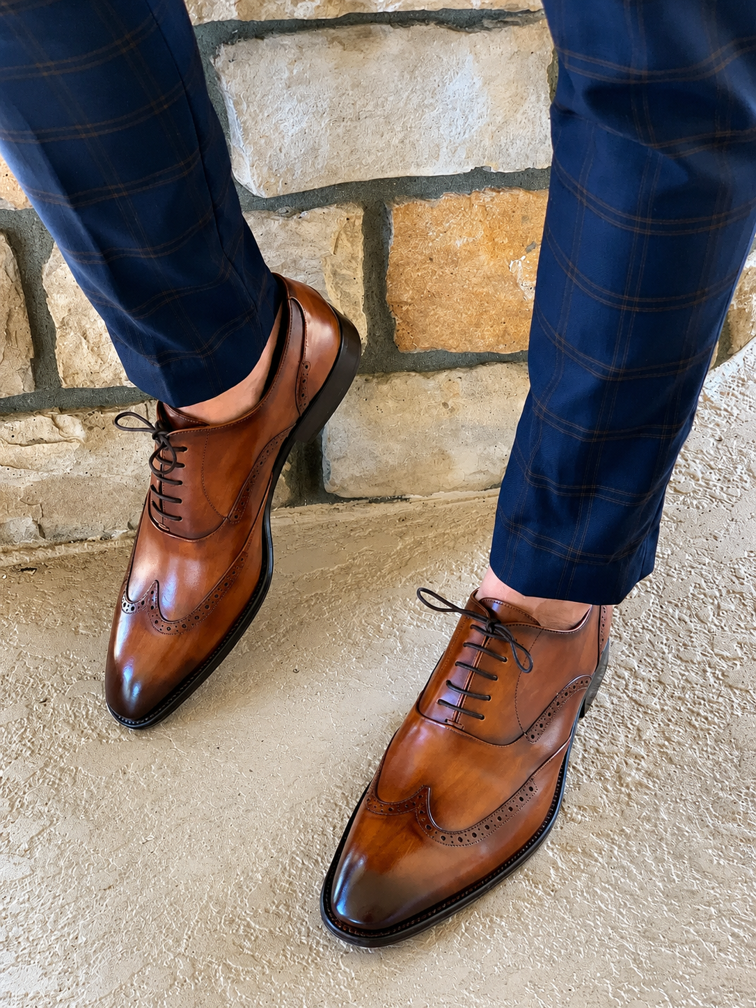 Brown leather dress shoes worn with blue pants against a stone wall background