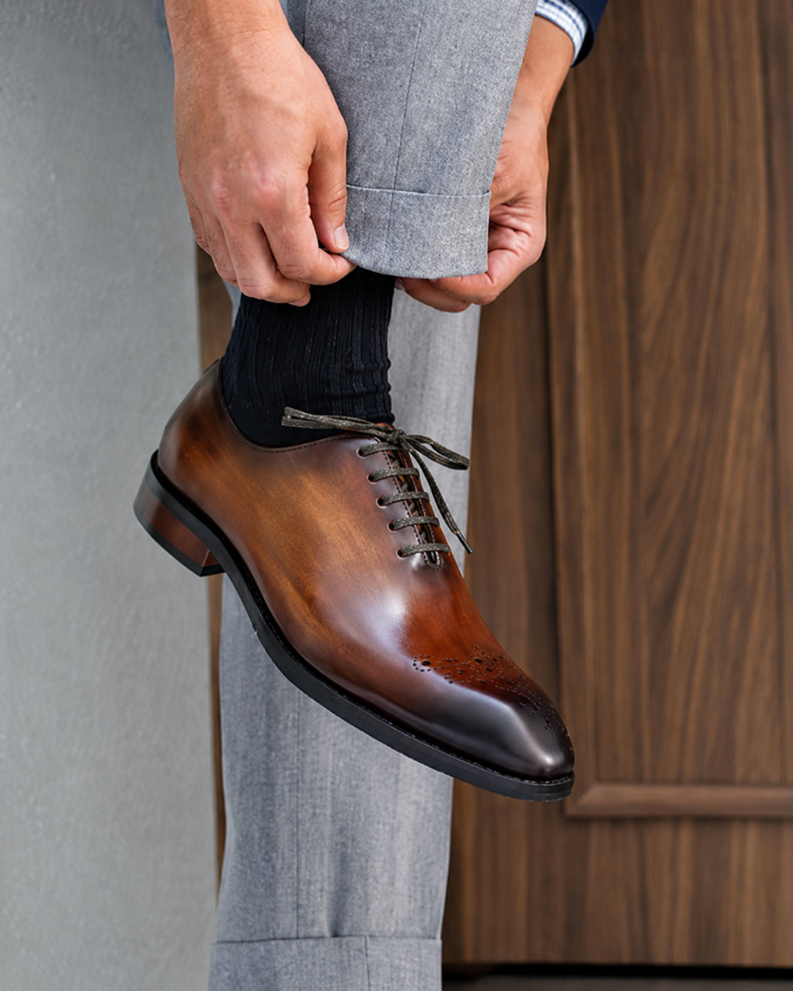 Brown dress shoe being worn with gray pants against a neutral background