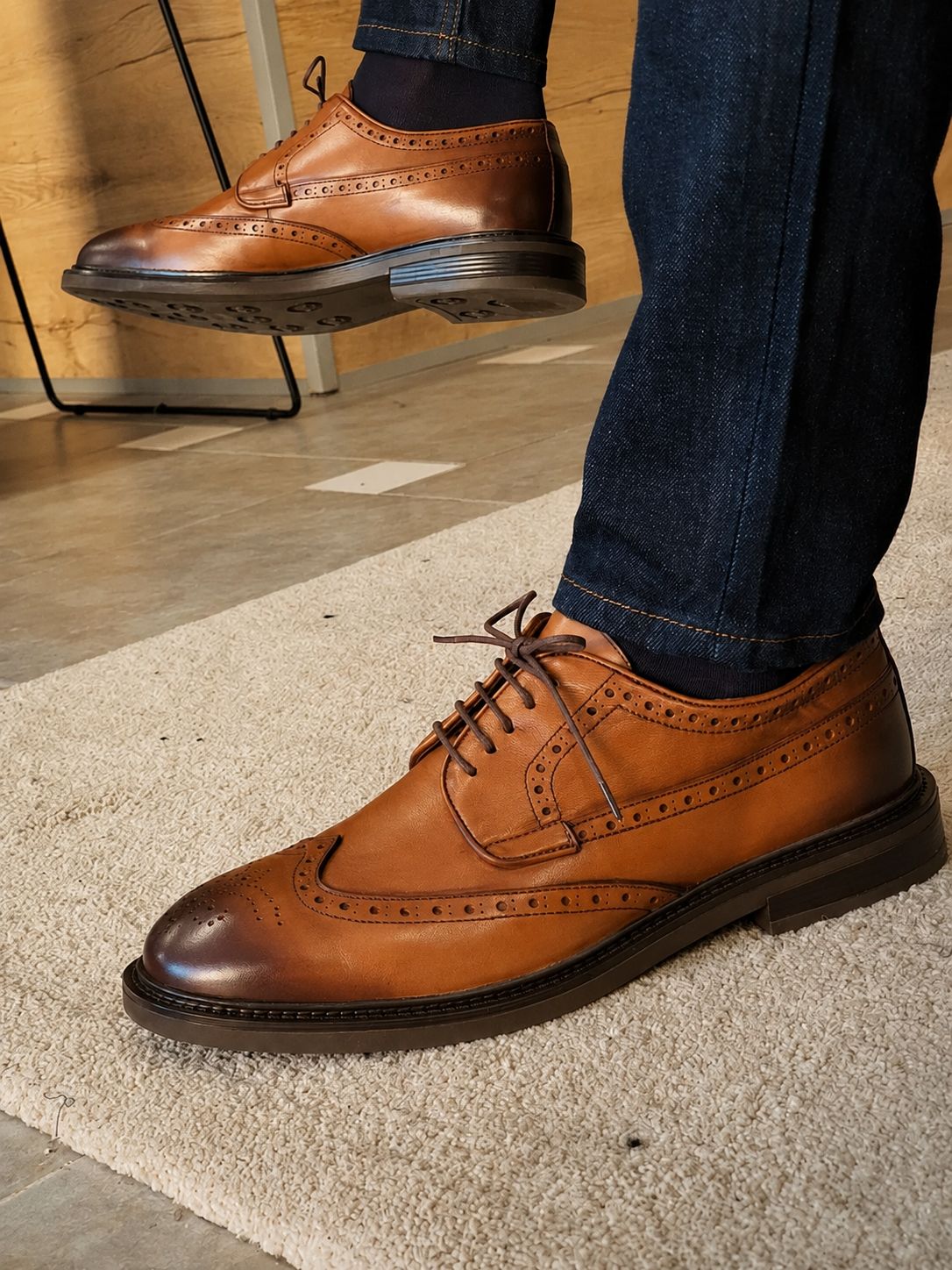 Brown leather shoes worn with dark jeans on a textured floor.