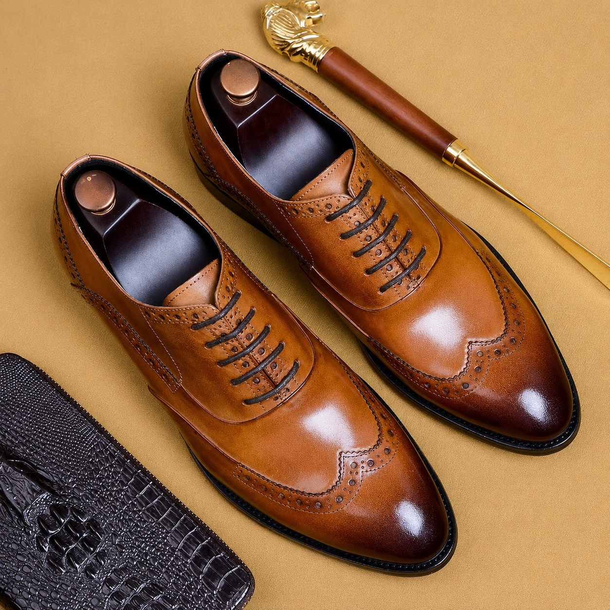 Premium tan leather wingtip Oxford brogue shoes with a polished finish, displayed in a flat lay on a beige background alongside a black textured wallet and a gold shoehorn.