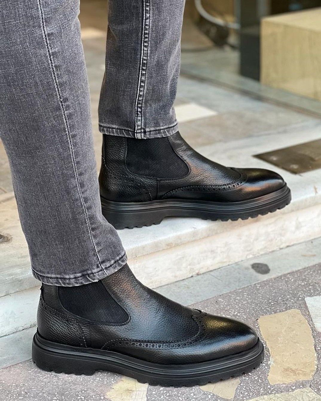 Black leather Chelsea boots worn with gray jeans on a sidewalk.