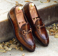 Pair of brown leather loafers with gold buckles on a concrete surface with leaves.
