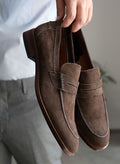 Brown suede loafers held by a person against a neutral background
