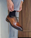 Brown dress shoe being worn with gray pants against a neutral background