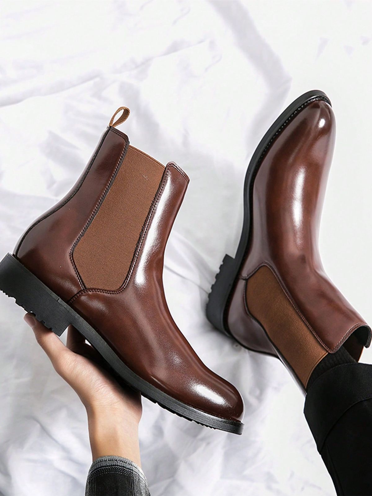 Brown leather Chelsea boots held by a hand on a white background