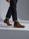 Brown suede Chelsea boots worn with black pants on a gray background