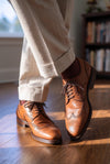 Brown dress shoes worn with beige pants on a wooden floor.
