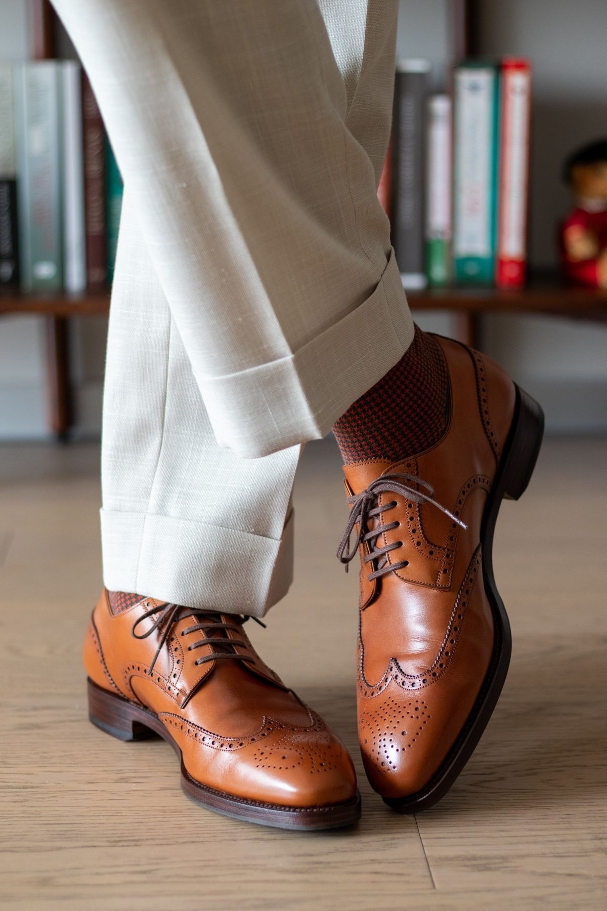 Brown leather dress shoes worn with white pants on a wooden floor.