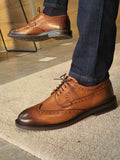 Brown leather shoes worn with blue jeans on a light wooden floor.