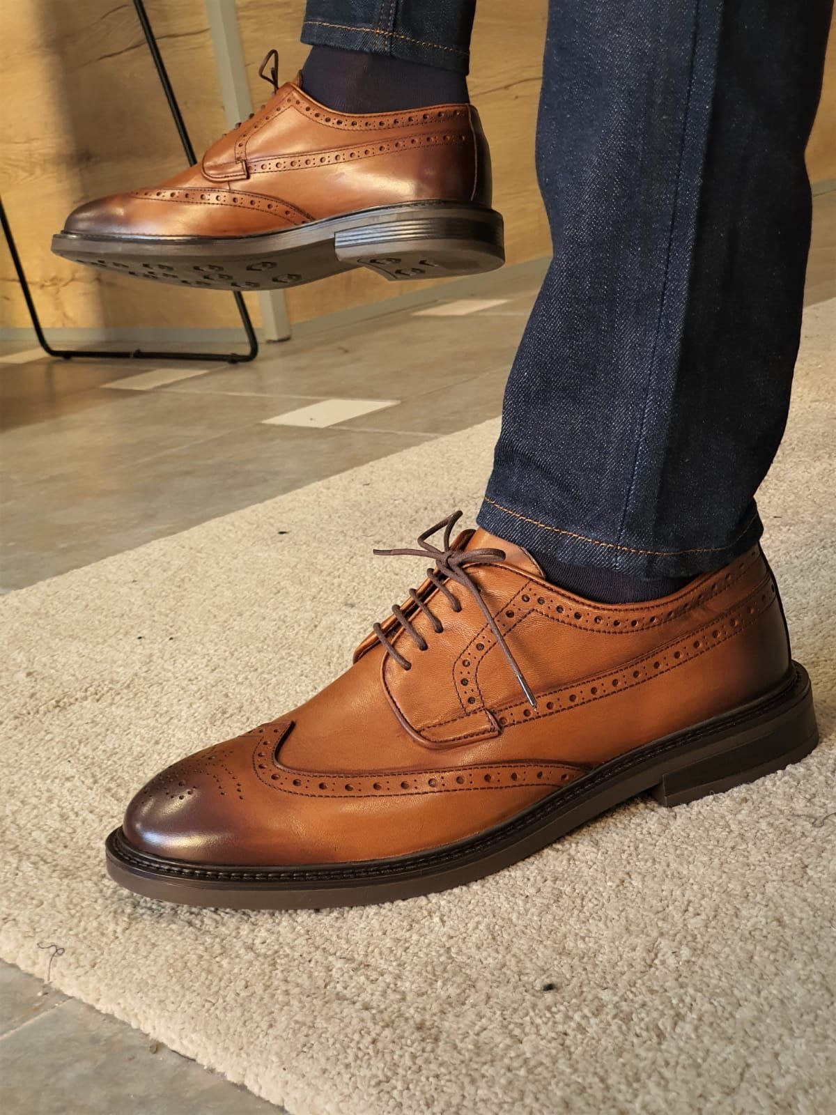 Brown leather shoes worn with blue jeans on a light wooden floor.