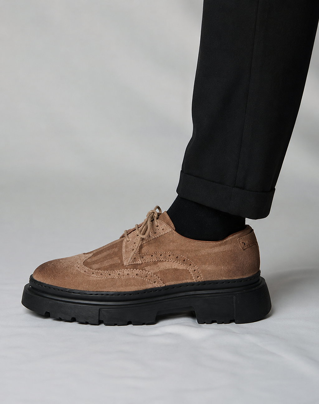 Brown suede brogue shoe with black sole worn by a person in black pants on a light gray background