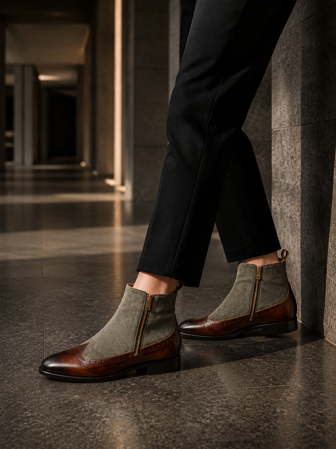 Gray and brown suede ankle boots worn with black pants on a dark floor.